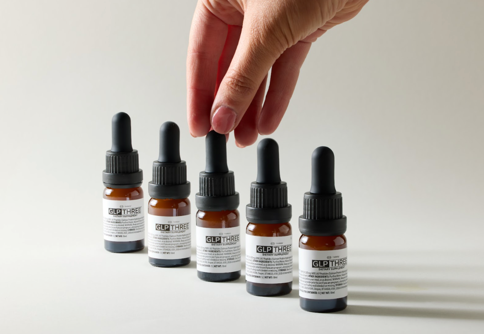 Five bottles of THREE GLP THREE dietary supplement arranged on a light surface as a hand reaches for one bottle. GLP THREE is formulated to support metabolic health and the body’s natural GLP-1-related processes.