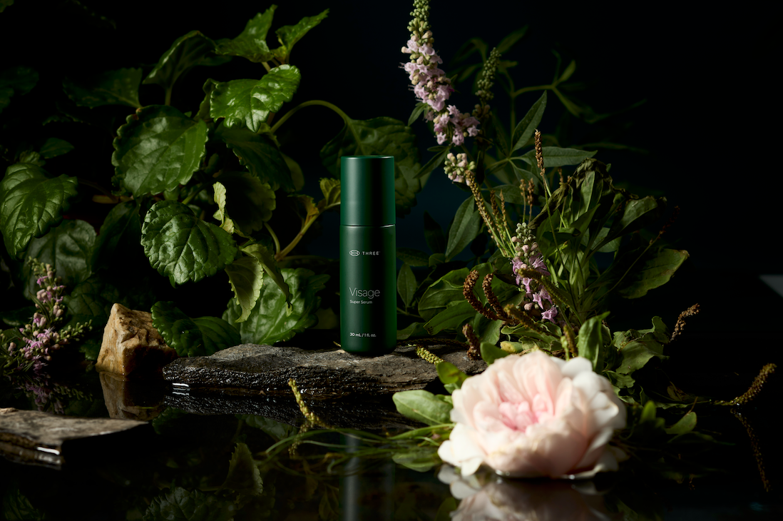 THREE VISAGE Super Serum displayed among lush green botanicals and delicate flowers. The neurocosmetic serum is formulated to support the skin-brain connection, hydration, and healthy-looking, radiant skin.