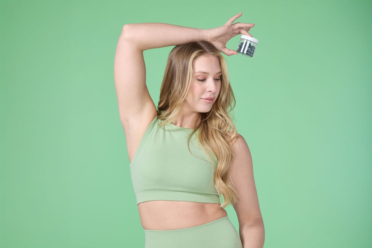 A woman in a green two piece holding up Purifí by THREE daily detox supplement