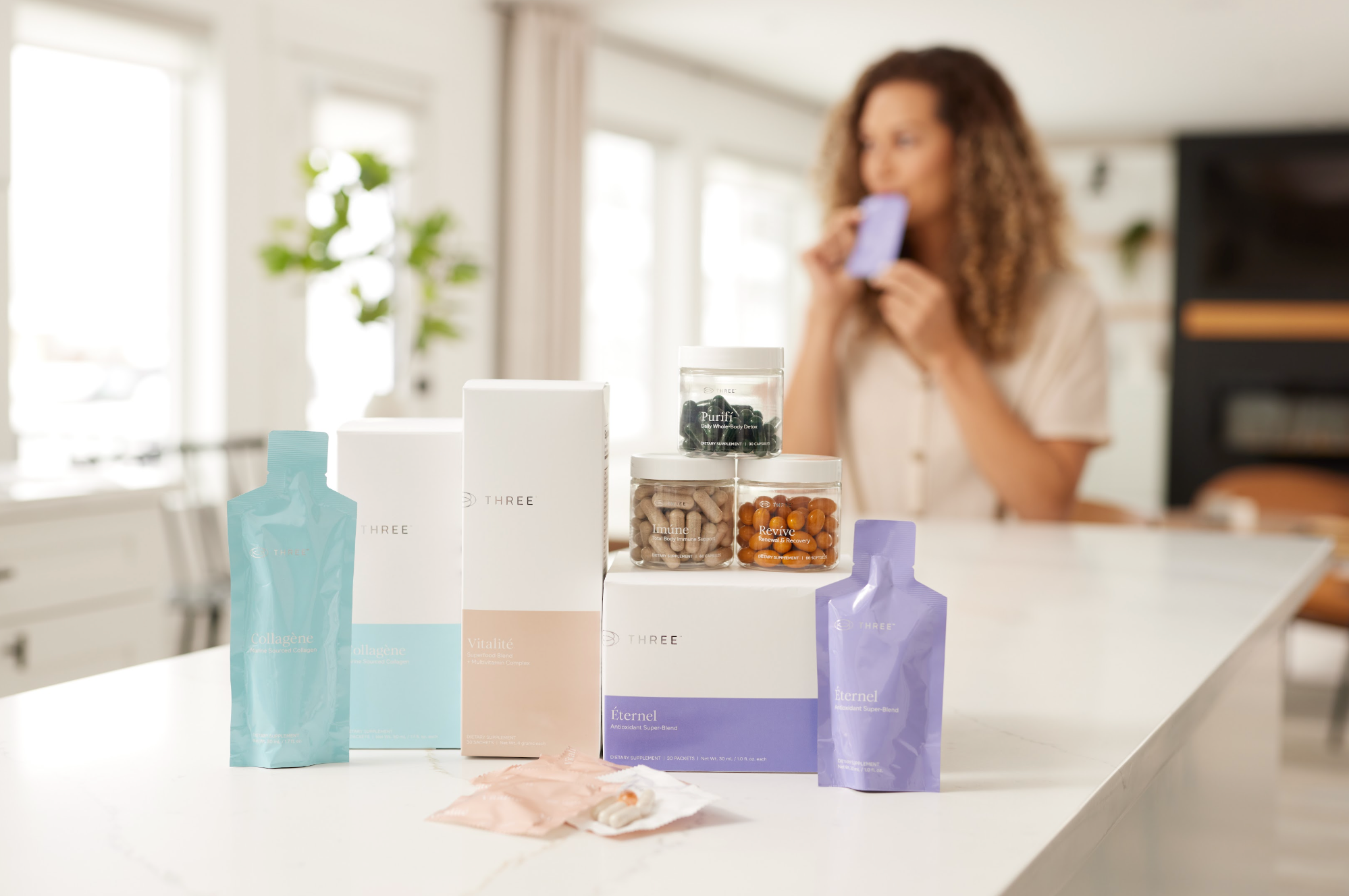 A collection of THREE International wellness products displayed on a kitchen counter, including Collagène, Vitalité, Éternel, Imúne, Revíve, and Purifí. The lifestyle setting highlights THREE’s science-driven approach to wellness and its diverse nutritional supplement portfolio.